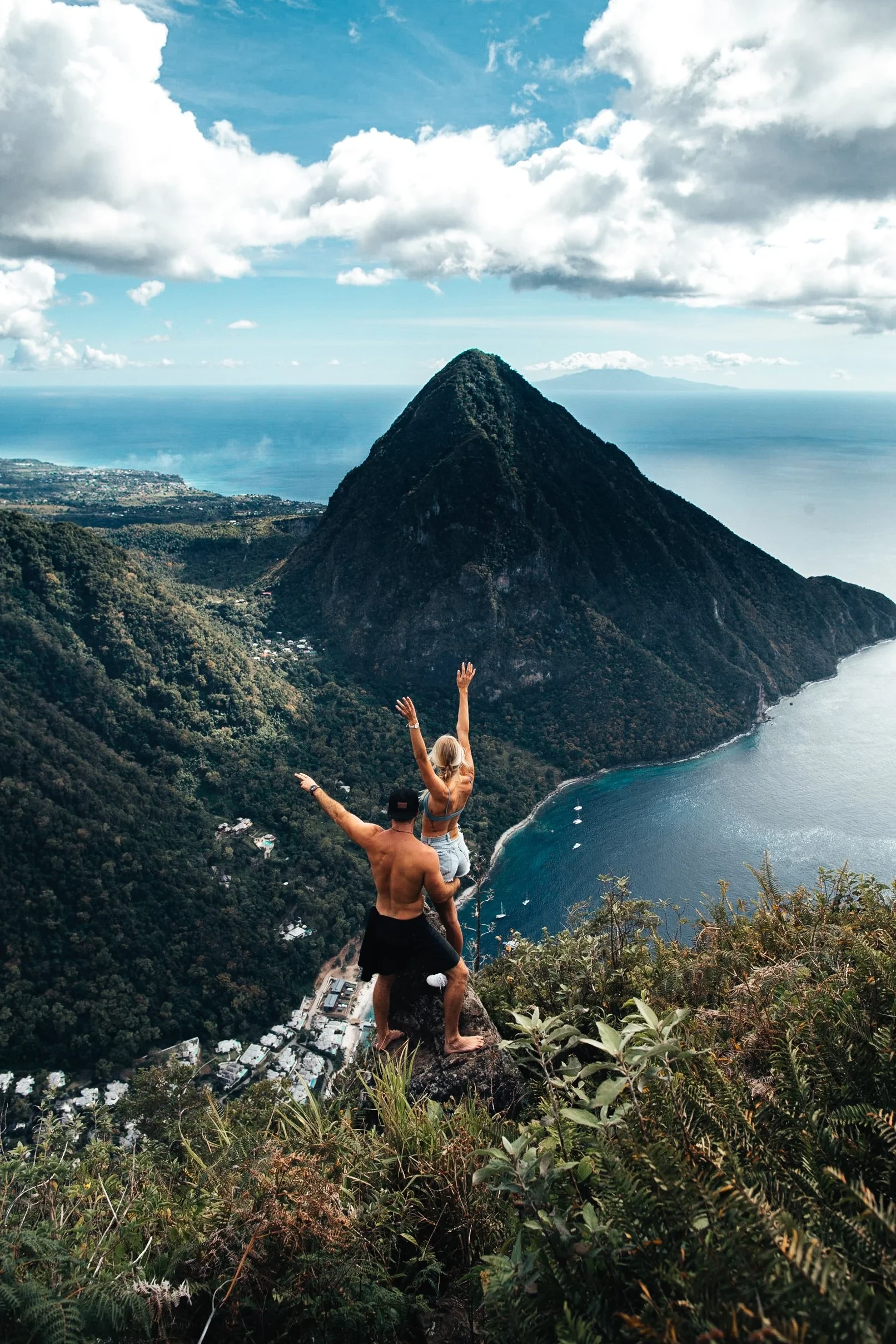 Two hikers celebrating with arms raised on the summit of Gros Piton with Petit Piton and the Caribbean Sea below